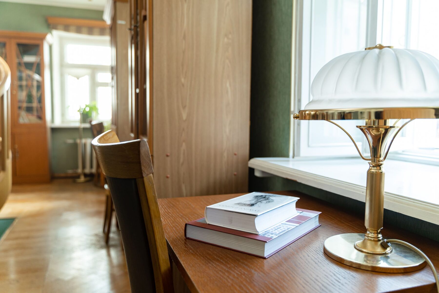 A brass desk lamp and a stack of books on a desk beside a window.